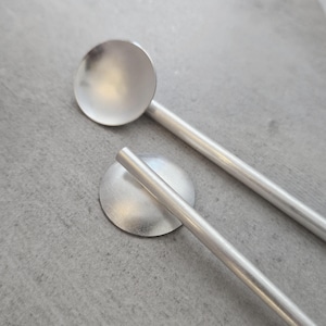 May include: Two silver spoons with rounded bowls and long, thin handles. The spoons are made of a smooth, shiny metal.