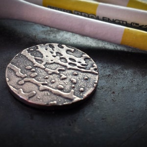 May include: A silver-coloured golf ball marker with a textured surface. The marker is circular in shape and has a raised design.