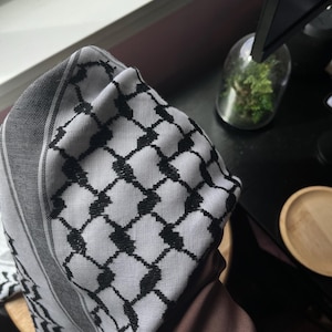 Könnte beinhalten: Ein schwarz-weiß karierter Keffiyeh-Schal. Der Schal hat ein sich wiederholendes geometrisches Muster aus schwarzen Quadraten auf weißem Grund. Die Kanten des Schals sind mit einem schwarz-grauen Streifen eingefasst.