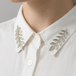 May include: Silver-toned leaf collar pins on a white button-down shirt. The pins are shaped like delicate branches with detailed leaves, adding a touch of nature-inspired elegance to the garment. The shirt's collar is crisp and clean.