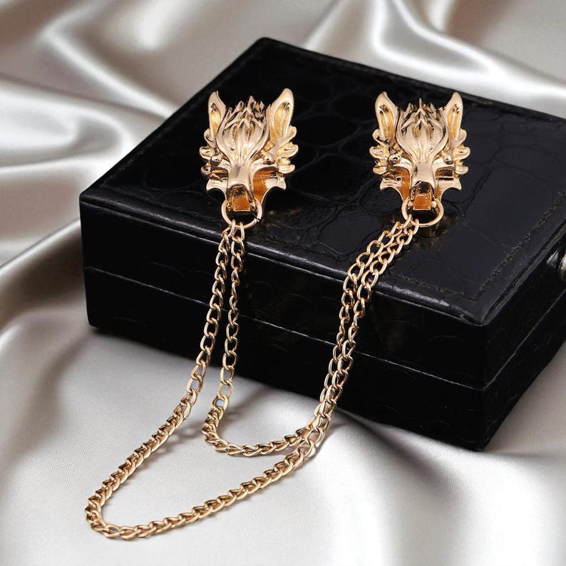 K&ouml;nnte beinhalten: Goldfarbene Wolfskopf-Reversnadeln mit Kettendetail. Die Anstecknadeln weisen detaillierte Wolfskopf-Designs auf und sind durch zarte goldene Ketten verbunden. Die Anstecknadeln werden auf einer schwarzen, strukturierten Box pr&auml;sentiert.