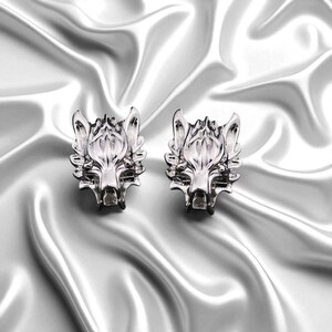 K&ouml;nnte beinhalten: Zwei silberfarbene Ohrstecker mit Wolfskopf. Die Ohrringe sind detailliert mit Merkmalen wie Schnauze, Ohren und Fell des Wolfes gestaltet. Sie werden auf einem gl&auml;nzenden, wei&szlig;en satinartigen Stoff pr&auml;sentiert.