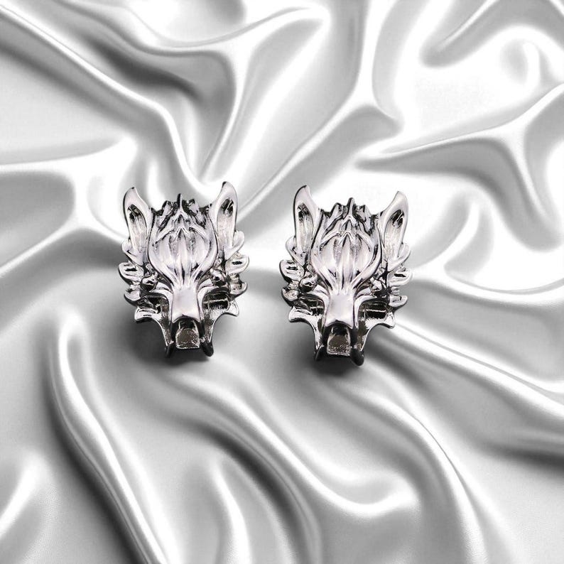 K&ouml;nnte beinhalten: Zwei silberfarbene Ohrstecker mit Wolfskopf. Die Ohrringe sind detailliert mit Merkmalen wie Schnauze, Ohren und Fell des Wolfes gestaltet. Sie werden auf einem gl&auml;nzenden, wei&szlig;en satinartigen Stoff pr&auml;sentiert.
