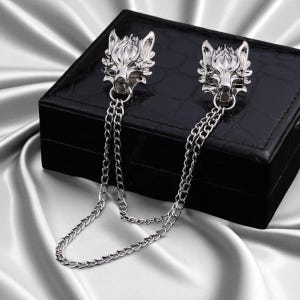 K&ouml;nnte beinhalten: Silberfarbene Metall-Kragenclips mit Wolfskopf-Designs. Jeder Clip hat einen detaillierten Wolfskopf und ist durch eine filigrane Kette verbunden. Die Clips werden auf einer schwarzen Box vor einem Satinhintergrund pr&auml;sentiert.