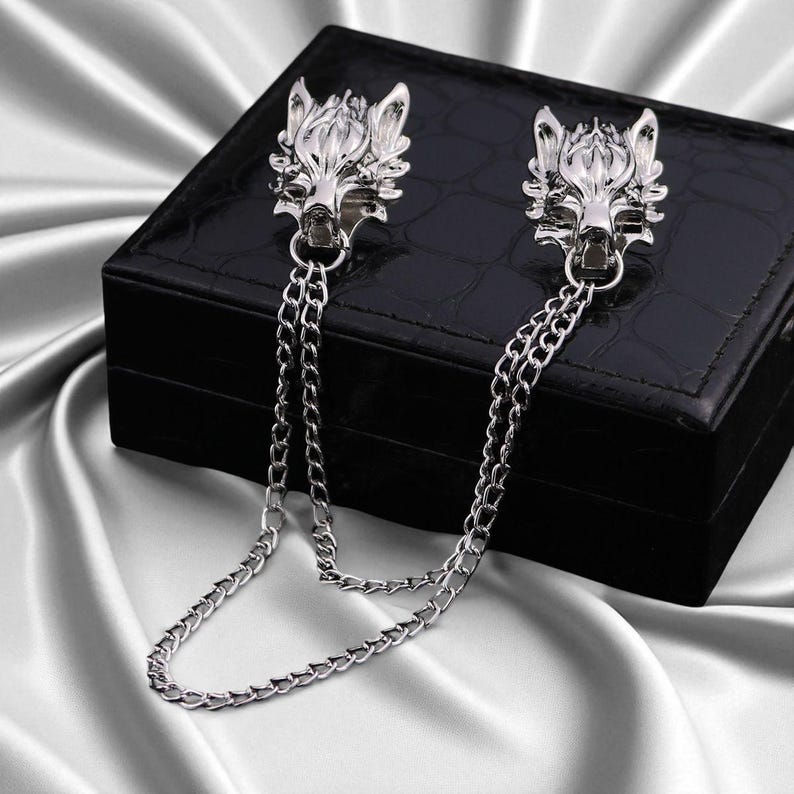K&ouml;nnte beinhalten: Silberfarbene Metall-Kragenclips mit Wolfskopf-Designs. Jeder Clip hat einen detaillierten Wolfskopf und ist durch eine filigrane Kette verbunden. Die Clips werden auf einer schwarzen Box vor einem Satinhintergrund pr&auml;sentiert.
