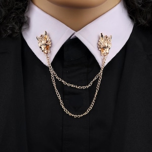 May include: Gold-colored collar clips featuring wolf head designs, connected by delicate chains. The clips are attached to a white collared shirt and black suit, creating a stylish accessory. This jewelry piece adds a unique touch to formal wear.