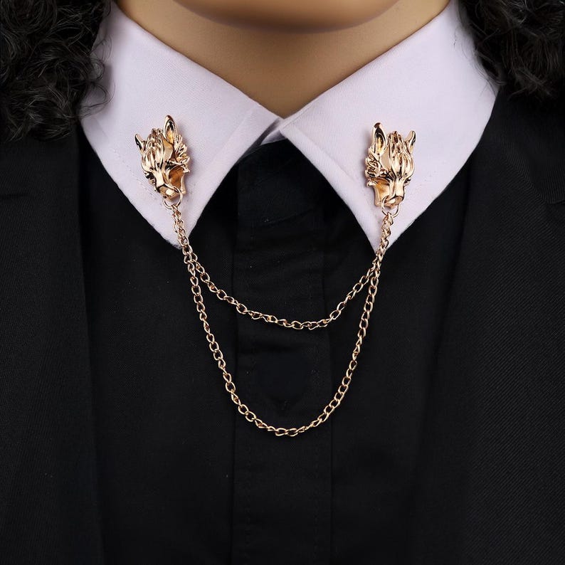 K&ouml;nnte beinhalten: Goldfarbene Kragenclips mit Wolfskopf-Design, verbunden durch filigrane Ketten. Die Clips sind an einem wei&szlig;en Hemdkragen und einem schwarzen Anzug befestigt, was ein stilvolles Accessoire ergibt. Dieses Schmuckst&uuml;ck verleiht formeller Kleidung eine einzigartige Note.