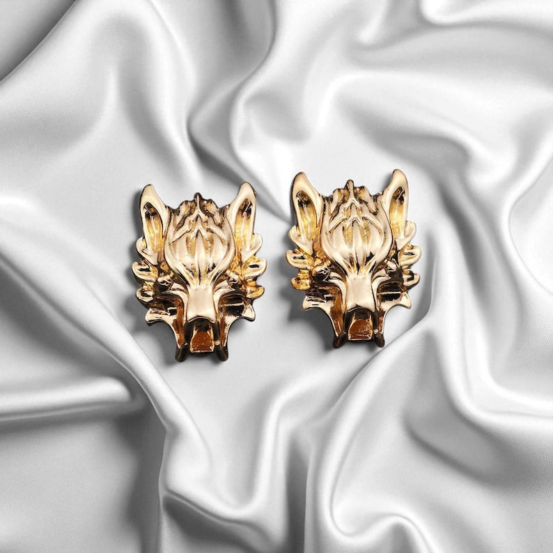 K&ouml;nnte beinhalten: Ein Paar goldfarbene Ohrstecker mit Wolfskopf-Motiv. Die Ohrringe sind mit strukturiertem Fell und Gesichtsz&uuml;gen versehen. Pr&auml;sentiert auf wei&szlig;em Satin-Hintergrund.