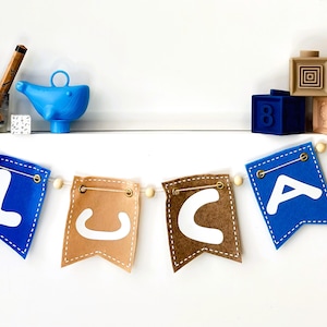 May include: A felt banner spelling "LICA" in white letters, with blue and tan pennants. Wooden beads string the pennants together. Other items include wooden blocks, a blue watering can, and pencils in a glass.