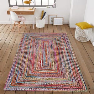 Peut inclure: Un tapis rectangulaire multicolore avec un motif rayé. Le tapis présente un motif répétitif de rectangles dans des tons de rouge, rose, bleu, jaune et vert. Le tapis est en matière tissée et est posé sur un sol en bois.