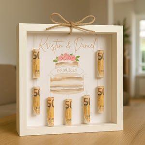 May include: A white framed shadow box with a wedding cake illustration and the names "Kristin & Daniel". Rolled Euro bills are suspended inside. A decorative twine bow is at the top. The date 09.09.2025 is printed on the cake.