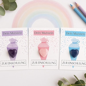 Op de afbeelding: Drie kaarten met illustraties van schooltüten, elk met een hartvormige steen. De tuiten zijn paars, roze en blauw, met bijpassende hartstenen. De kaarten tonen de tekst "Dein Mutstein" en "ZUR EINSCHULUNG". Een regenboog en potloden zijn op de achtergrond.