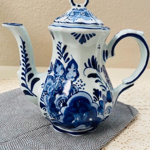 May include: A white ceramic teapot featuring intricate blue floral patterns and foliage designs. The teapot has a curved handle, a long spout, and a matching lid with a decorative finial. The surface is glossy with a smooth, rounded body shape.