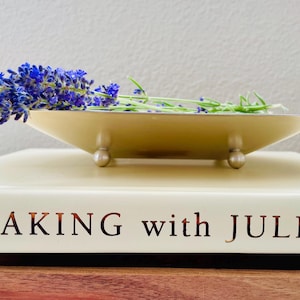 May include: A square, cream-colored metal tray with four small ball feet, holding a sprig of purple lavender. The tray rests on a thick, cream-colored book with the text "BAKING with JULIA" printed in dark, metallic lettering on the spine.