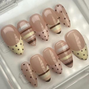 May include: A set of ten artificial nails in a clear plastic case. The nails feature a nude base with various designs, including polka dots, stripes, and a French tip with yellow polka dots. The nails are oval-shaped.