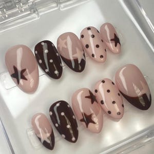 May include: A set of oval-shaped false nails in a clear plastic case. The nails feature a variety of designs, including solid brown, polka dots, and star patterns. The color palette includes shades of brown and pink.