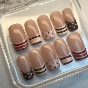 May include: A set of ten artificial nails in a clear plastic case. The nails have a nude base with various designs, including polka dots, stripes in burgundy, white, and brown, and a solid brown tip with white dots. The nails are oval-shaped.