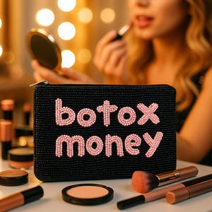 Puede incluir: Bolsa de cosméticos negra con cuentas con las palabras "botox money" en cuentas rosas. Pinceles de maquillaje, polveras y barras de labios están dispuestos alrededor de la bolsa. El fondo está borroso, con una persona aplicándose lápiz labial.