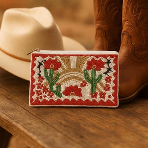 May include: A beaded clutch with a Southwestern design featuring a sun, cacti, and flowers in red, gold, green, and white. The clutch has a white zipper and is set on a wooden surface with a cowboy hat and boots in the background.