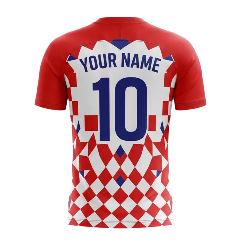 Pu&ograve; includere: Maglia da calcio rossa e bianca con la scritta "YOUR NAME" sopra il numero "10" in blu. La maglia presenta un motivo a scacchi nella parte inferiore e maniche rosse.