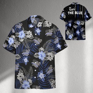 Puede incluir: Camisa negra de manga corta con un estampado floral y de bandera. El diseño incluye flores de hibisco azules, flores blancas y banderas americanas. La parte trasera de la camisa tiene el texto "Back the Blue".