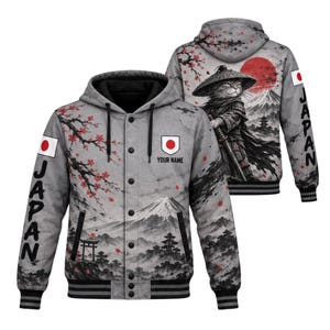 Puede incluir: Una sudadera gris con botones y capucha que presenta un gráfico de un gato samurái, el monte Fuji y flores de cerezo. Las mangas muestran la palabra JAPAN y la bandera japonesa. El pecho incluye un emblema con la bandera y el texto YOUR NAME.
