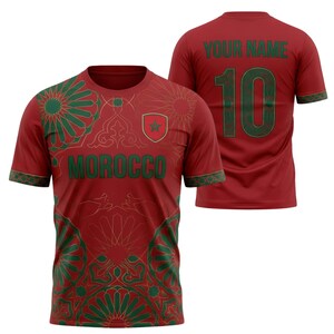 May include: Red soccer jersey with green and gold geometric patterns and the word "MOROCCO" across the chest. The back of the jersey has the number 10 and the text "YOUR NAME" in green.