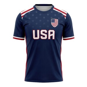 May include: A navy blue athletic jersey with the word "USA" in white block letters across the chest. The jersey features a small American flag emblem and a pattern of white stars. Red and white stripes accent the shoulders and sleeves.