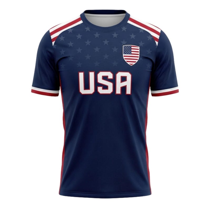 May include: A navy blue athletic jersey with the word "USA" in white block letters across the chest. The jersey features a small American flag emblem and a pattern of white stars. Red and white stripes accent the shoulders and sleeves.