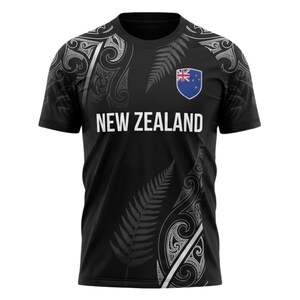 May include: Black t-shirt with white text that reads "NEW ZEALAND". The shirt features a fern design and a New Zealand flag emblem. The shirt has a crew neck and short sleeves.