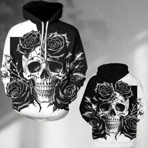 Puede incluir: Sudadera con capucha en blanco y negro con un diseño de calavera rodeada de rosas negras. La sudadera tiene un diseño dividido con una combinación de colores en blanco y negro. La calavera está detallada y las rosas añaden una estética gótica.