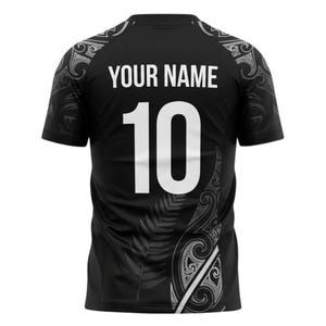 May include: Black athletic jersey with the text "YOUR NAME" above the number "10" in white. The jersey has decorative white patterns on the shoulders and sides, with a fern design. The jersey is short-sleeved.