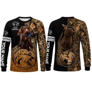 May include: A long-sleeve shirt featuring a brown hunting dog holding a duck in its mouth, surrounded by marsh grass and reeds. The design includes the text "DUCK HUNTING" and "YOUR NAME" on a black and camouflage background with flying ducks.