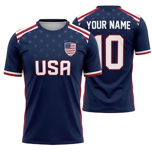 May include: A navy blue soccer jersey with white and red accents. The front features the word "USA" in white block letters, the American flag, and a pattern of stars. The back displays the number 10 and the text "YOUR NAME" in white.