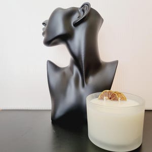 Puede incluir: Un busto negro de cabeza y hombros de mujer se encuentra junto a una vela blanca en un recipiente de vidrio esmerilado. La vela est&aacute; cubierta con dos rodajas de c&iacute;tricos secos. El busto es liso y brillante.