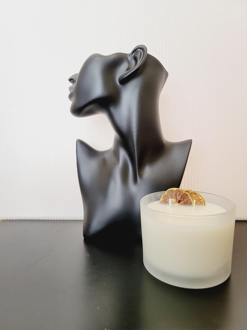 Puede incluir: Un busto negro de cabeza y hombros de mujer se encuentra junto a una vela blanca en un recipiente de vidrio esmerilado. La vela est&aacute; cubierta con dos rodajas de c&iacute;tricos secos. El busto es liso y brillante.