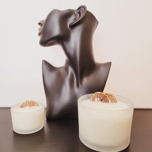 Puede incluir: Un busto negro de la cabeza y los hombros de una mujer se encuentra detr&aacute;s de dos velas blancas en recipientes de vidrio esmerilado. Cada vela est&aacute; rematada con una rodaja de naranja seca. Las velas est&aacute;n sobre una superficie oscura.