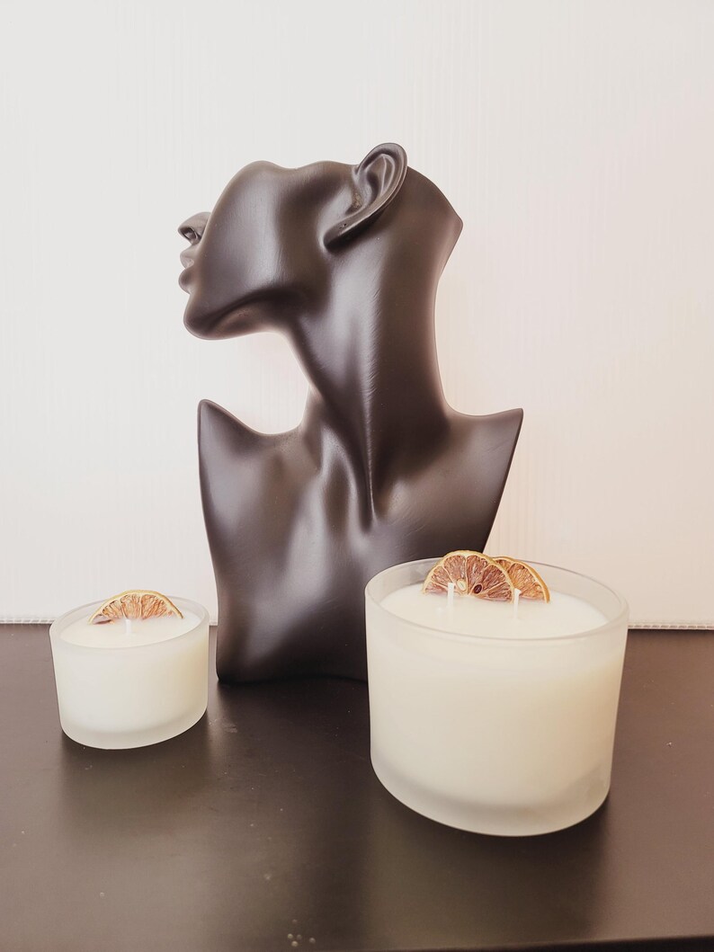 Puede incluir: Un busto negro de la cabeza y los hombros de una mujer se encuentra detr&aacute;s de dos velas blancas en recipientes de vidrio esmerilado. Cada vela est&aacute; rematada con una rodaja de naranja seca. Las velas est&aacute;n sobre una superficie oscura.