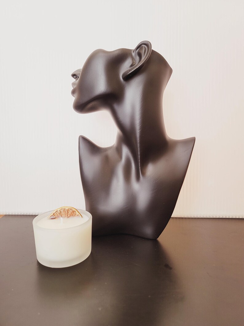 Puede incluir: Un busto de maniqu&iacute; negro con un acabado liso se encuentra junto a un peque&ntilde;o portavelas de cristal esmerilado. La vela es blanca y est&aacute; rematada con una rodaja de c&iacute;trico seco. El busto es un art&iacute;culo decorativo.