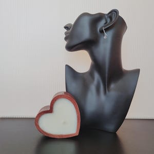 Puede incluir: Una vela en forma de coraz&oacute;n con un marco de madera marr&oacute;n rojizo y cera blanca. Una cabeza de maniqu&iacute; negra con un pendiente plateado est&aacute; en el fondo. La vela est&aacute; sobre una superficie oscura.
