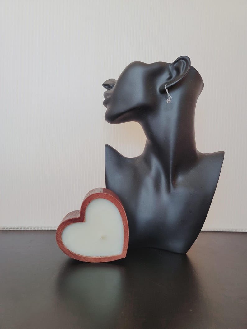 Puede incluir: Una vela en forma de coraz&oacute;n con un marco de madera marr&oacute;n rojizo y cera blanca. Una cabeza de maniqu&iacute; negra con un pendiente plateado est&aacute; en el fondo. La vela est&aacute; sobre una superficie oscura.