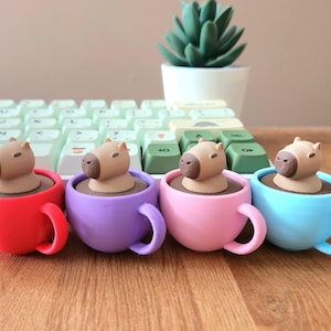 May include: Four colorful coffee cups, red, purple, pink, and blue, each holding a small capybara figurine. The figurines are light brown and appear to be sitting in a dark brown liquid. A keyboard and a small potted succulent are in the background.