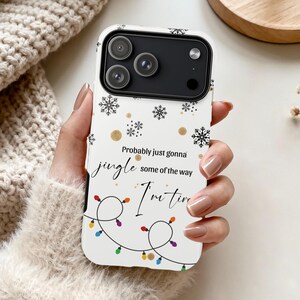 May include: White phone case with a festive design. The case features black snowflakes, gold dots, and a string of colorful lights. Text on the case reads "Probably just gonna jingle some of the way I'm tired."