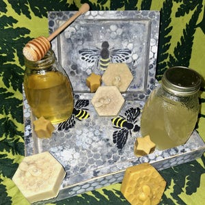 Puede incluir: Una bandeja de madera con un patrón de panal de abeja contiene dos frascos de vidrio con miel, un cucharón de miel de madera y jabones con temática de abejas. Los jabones tienen forma de estrella y hexágono, con detalles de abeja y panal. El fondo es un patrón floral verde.