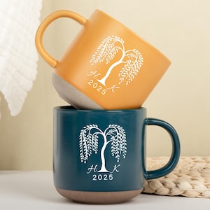 Puede incluir: Dos tazas de cerámica, una mostaza y otra verde azulado, apiladas. Cada taza presenta un diseño de sauce llorón blanco con las iniciales "H K" y el año "2025". Las tazas tienen una base gris claro y un asa curva.
