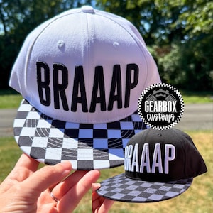 May include: Two baseball caps, one white and one black, with the word "BRAAAP" embroidered on the front. The white cap has a black and white checkered brim. The black cap has a gray and black checkered brim. A Gearbox Customs logo is also visible.