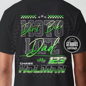 Pode incluir: Camiseta preta com um design gráfico verde e branco. O design inclui as palavras "Dirt Bike Life Dad" e o nome "Chase Holman" com o número "123". O logotipo da Kawasaki também é visível.