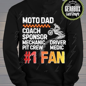 Peut inclure: Un sweat à capuche noir avec du texte blanc qui dit "MOTO DAD COACH SPONSOR MECHANIC DRIVER PIT CREW MEDIC #1 FAN". Un graphique d'un pilote de motocross et le logo Gearbox Customs sont également présents.
