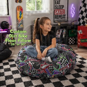 May include: A round floor pillow with a dirt bike theme, featuring a colorful design with motorcycles, checkered flags, and the text "BRAAAP". The pillow is available in 26 or 30-inch sizes. A young girl is sitting on the pillow.