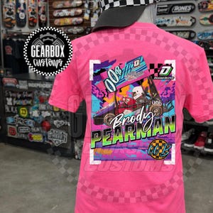 Può includere: Una maglietta rosa con un motivo a scacchi e una grafica di un'auto da corsa con il nome "Brody Pearman" e il numero "006". La maglietta presenta anche il testo "Gearbox Customs".